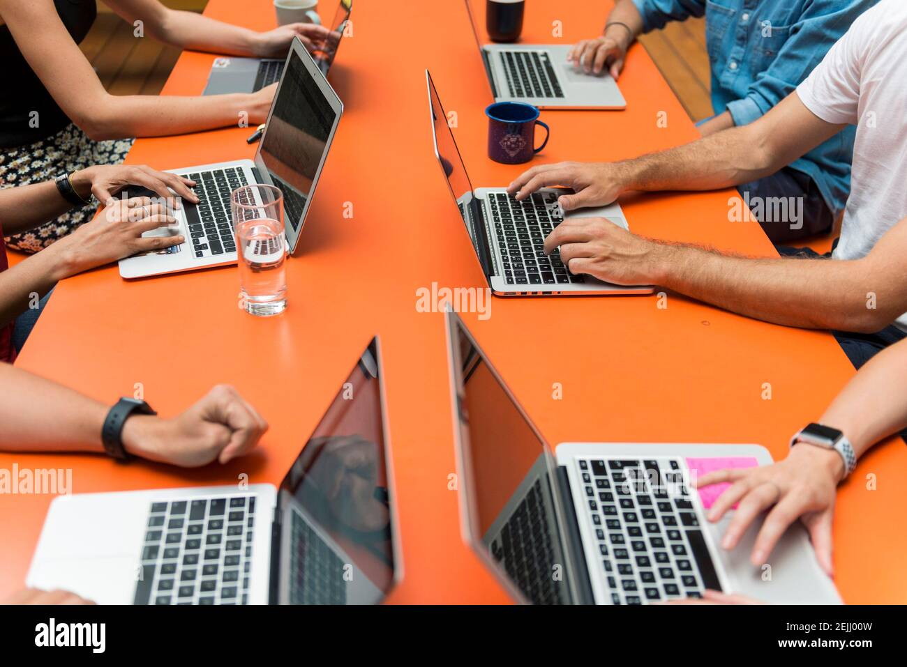 Six people sat around a bright orange table in a meeting while working on the laptop computers Stock Photo