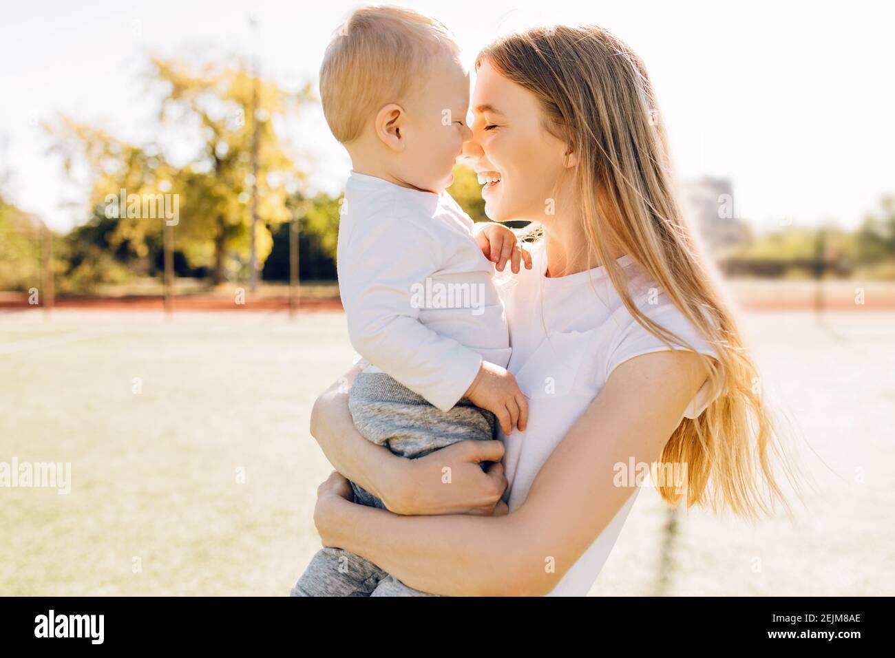 Caring young mother with baby, loving mom kissing hugs little child enjoying tender family moment outdoors on a sunny day, Mother's Day Stock Photo