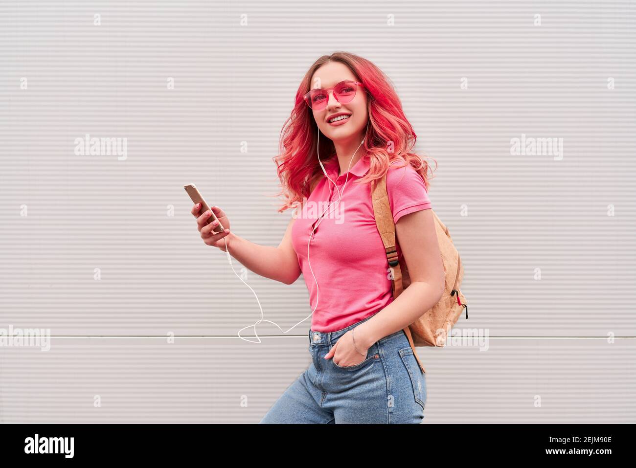 Young blogger woman with pink dyed hairs staying near the grey wall and using smartphone Stock Photo