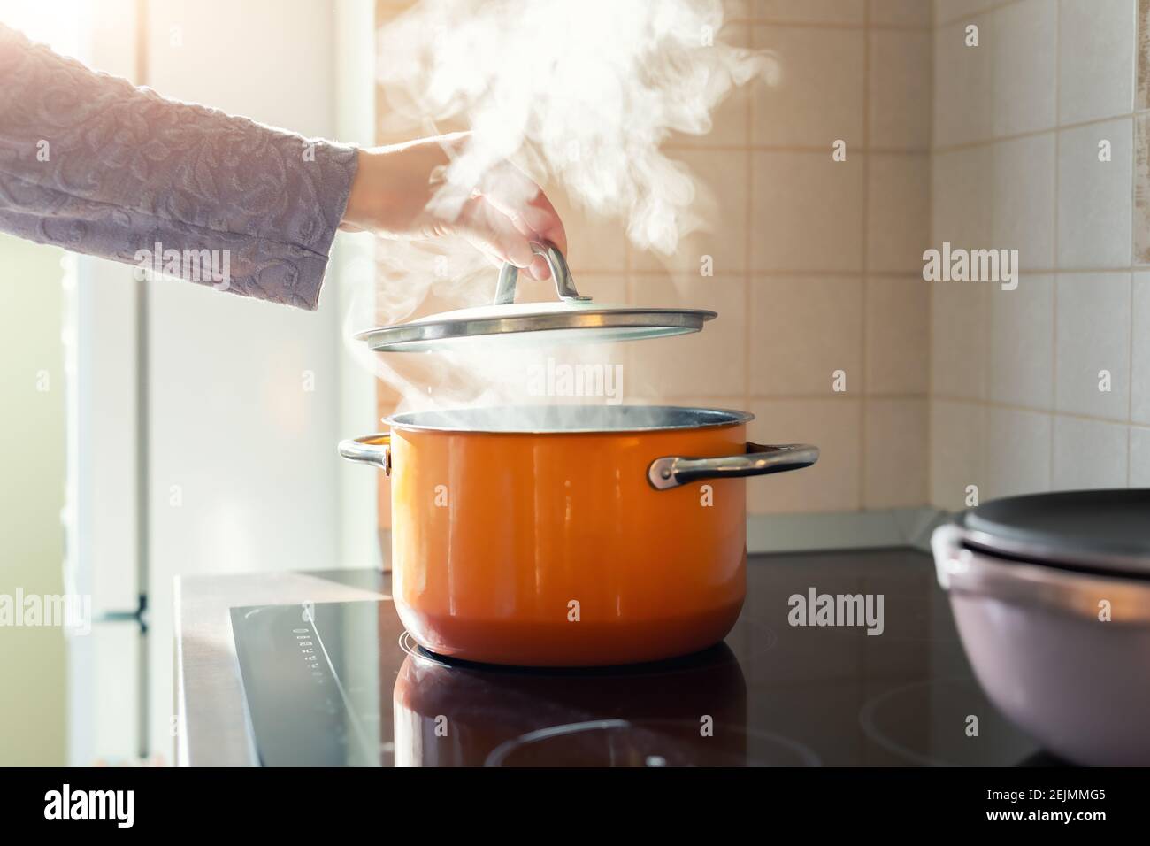 Female hand open lid of enamel steel cooking pan on electric hob with boiling water or soup and