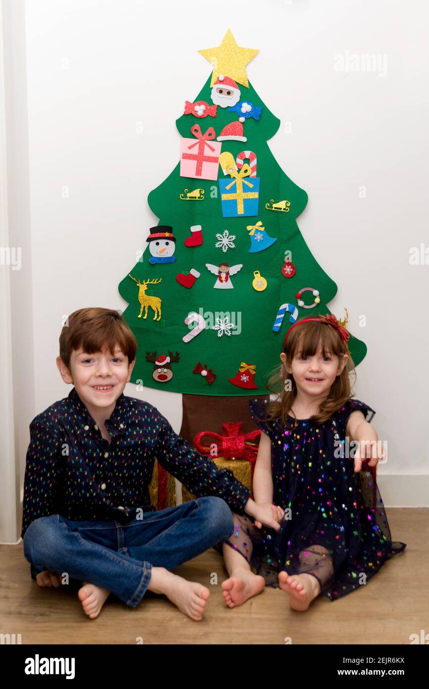A cute, brown-haired, blue-eyed baby girl and a cute, red-haired, blue-eyed boy smartly dressed sitting on the floor in front of a Christmas tree Stock Photo