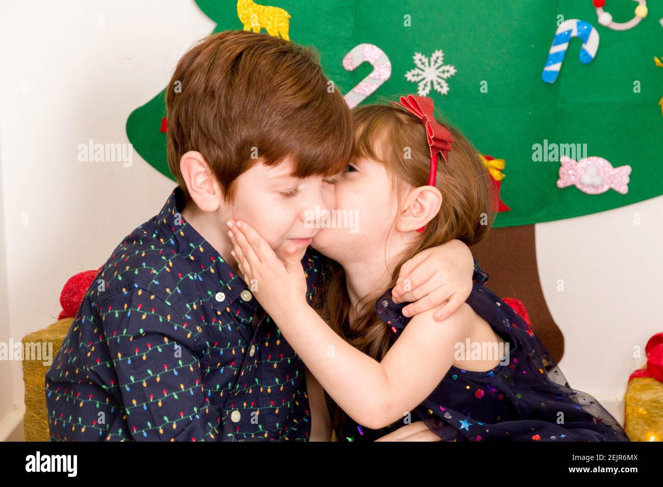 A cute, brown-haired, blue-eyed baby girl and a cute, red-haired, blue-eyed boy smartly dressed kissing on front of a Christmas tree Stock Photo