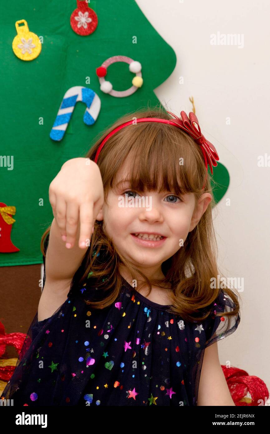 A  cute, brown-haired, blue-eyed baby smartly dressed smiling in front of a Christmas tree with her hand up Stock Photo