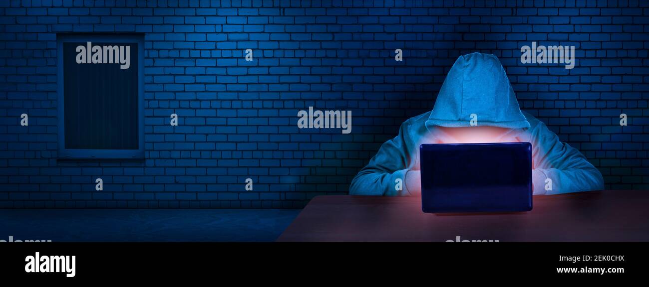 The hacker works behind a laptop screen in a room with a brick wall. Internet crime. The concept of hacking and malware, cybersecurity. Digital crimes Stock Photo