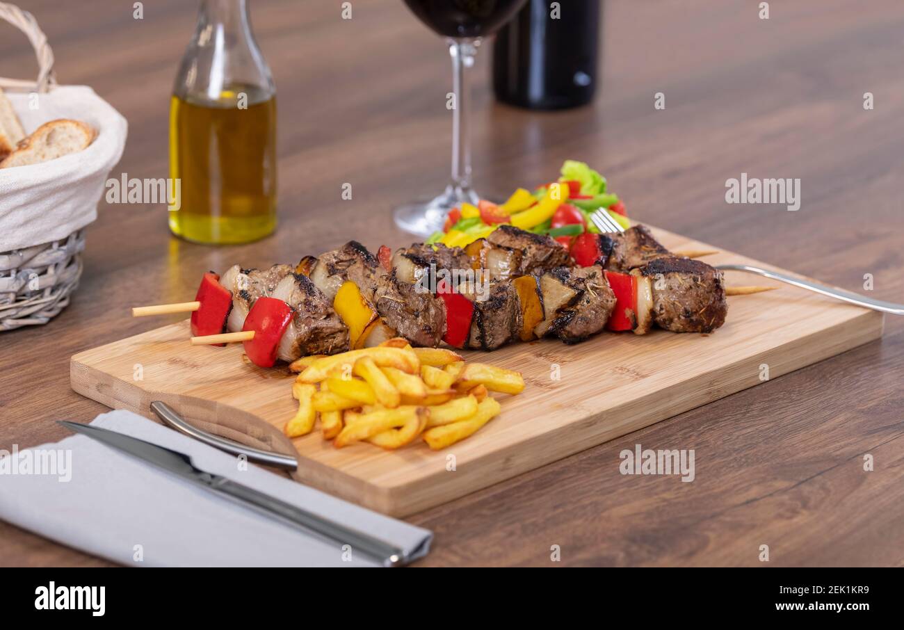 Appetizing beef skewers surrounded by out of focus french fries and cut vegetables on a wooden board. Selective focus. Stock Photo