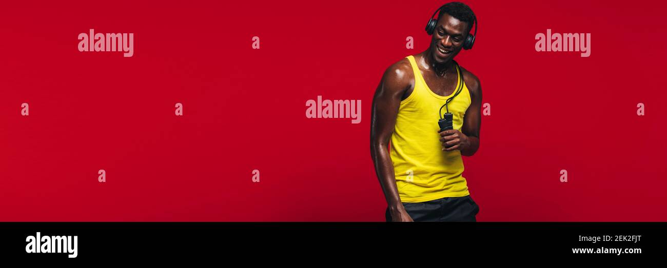 African muscular man with jumping rope listening to music on headphones. Fit young man relaxing after workout on red background with copy space. Stock Photo