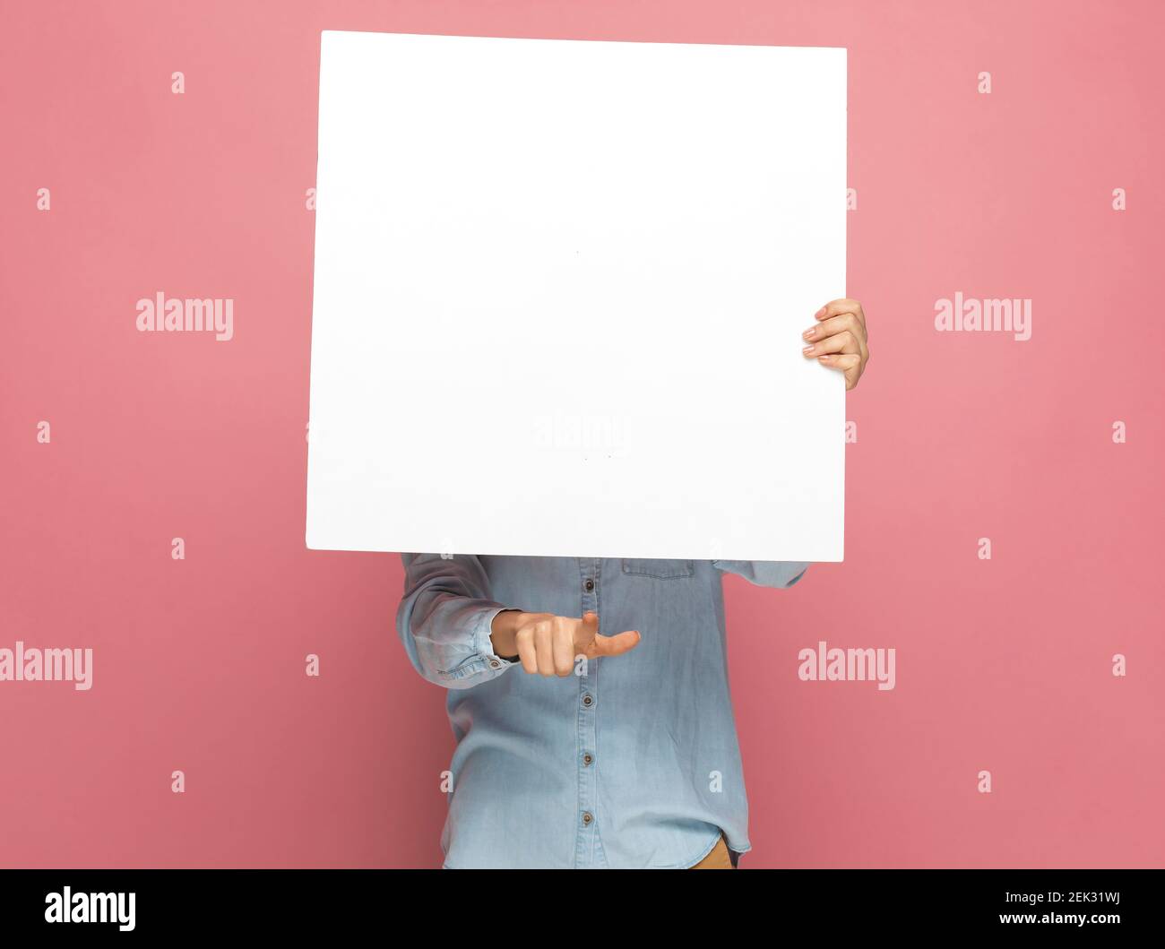 casual woman pointing at the camera behind a blank billboard against pink background Stock Photo