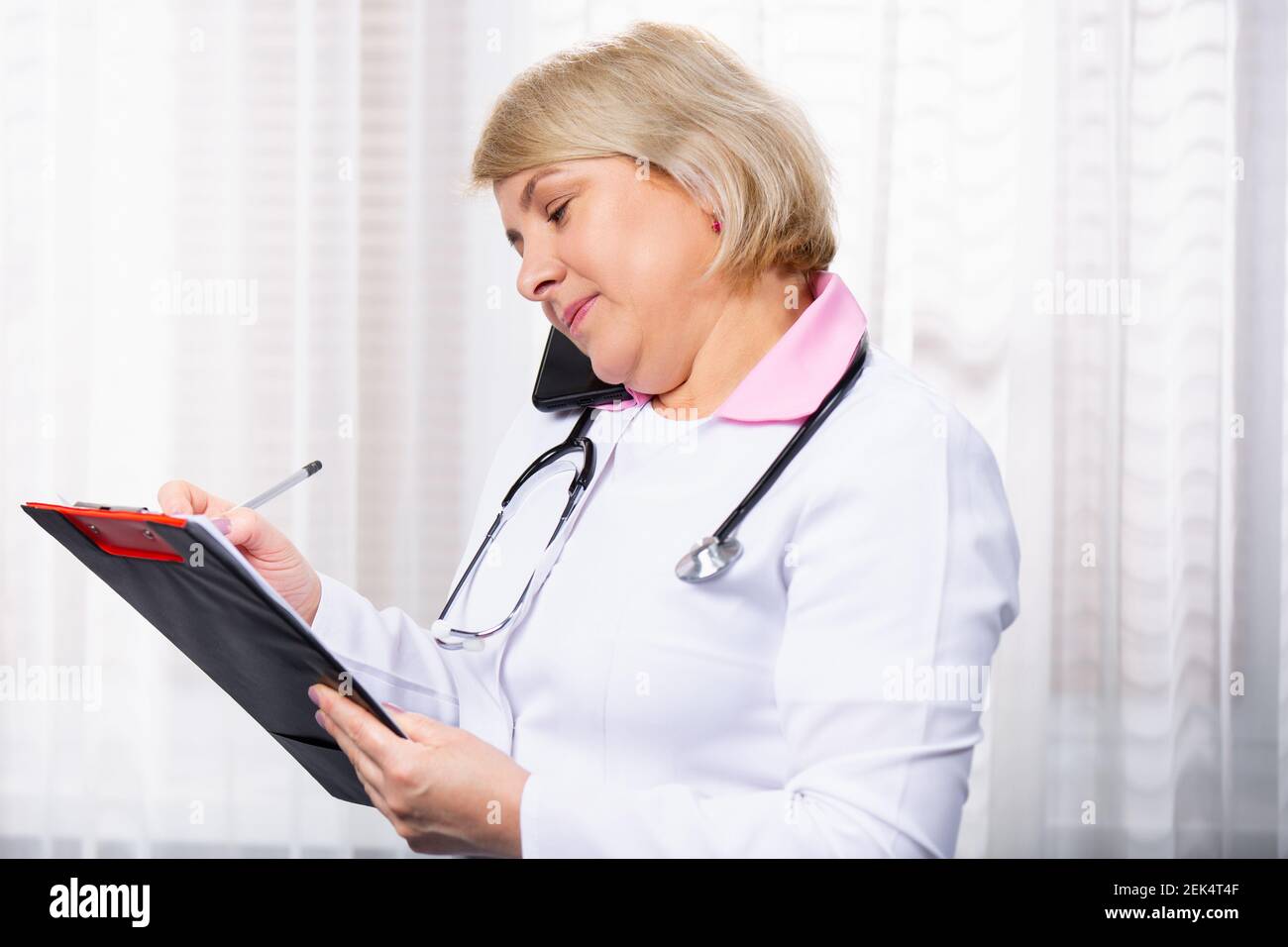 Caucasian doctor handsome woman with stethoscope talking on mobile phone, smartphone calls at clinic, checking healthcare of customer on paper Stock Photo