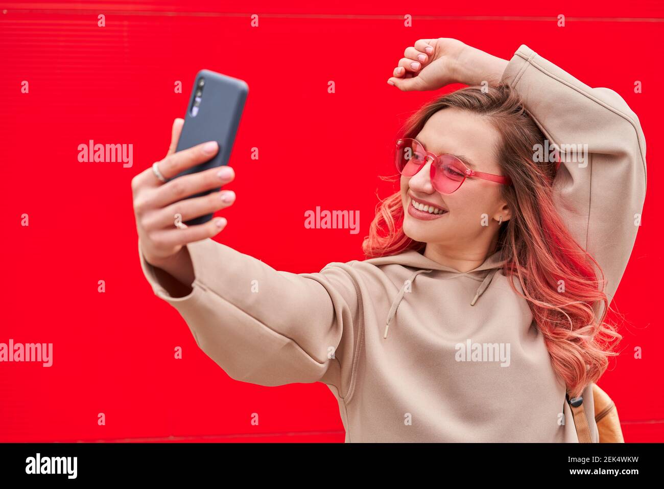 Young blogger woman with pink dyed hairs staying near the red wall and making selfie by smartphone Stock Photo