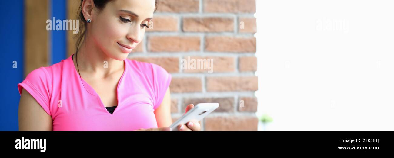Beautiful woman in pink T-shirt stand in fitness room and hold phone in her hands. Stock Photo