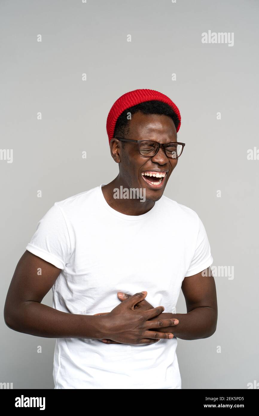 Overjoyed Black millennial man shows her broad smile, holding his stomach from laughter. Studio. Stock Photo