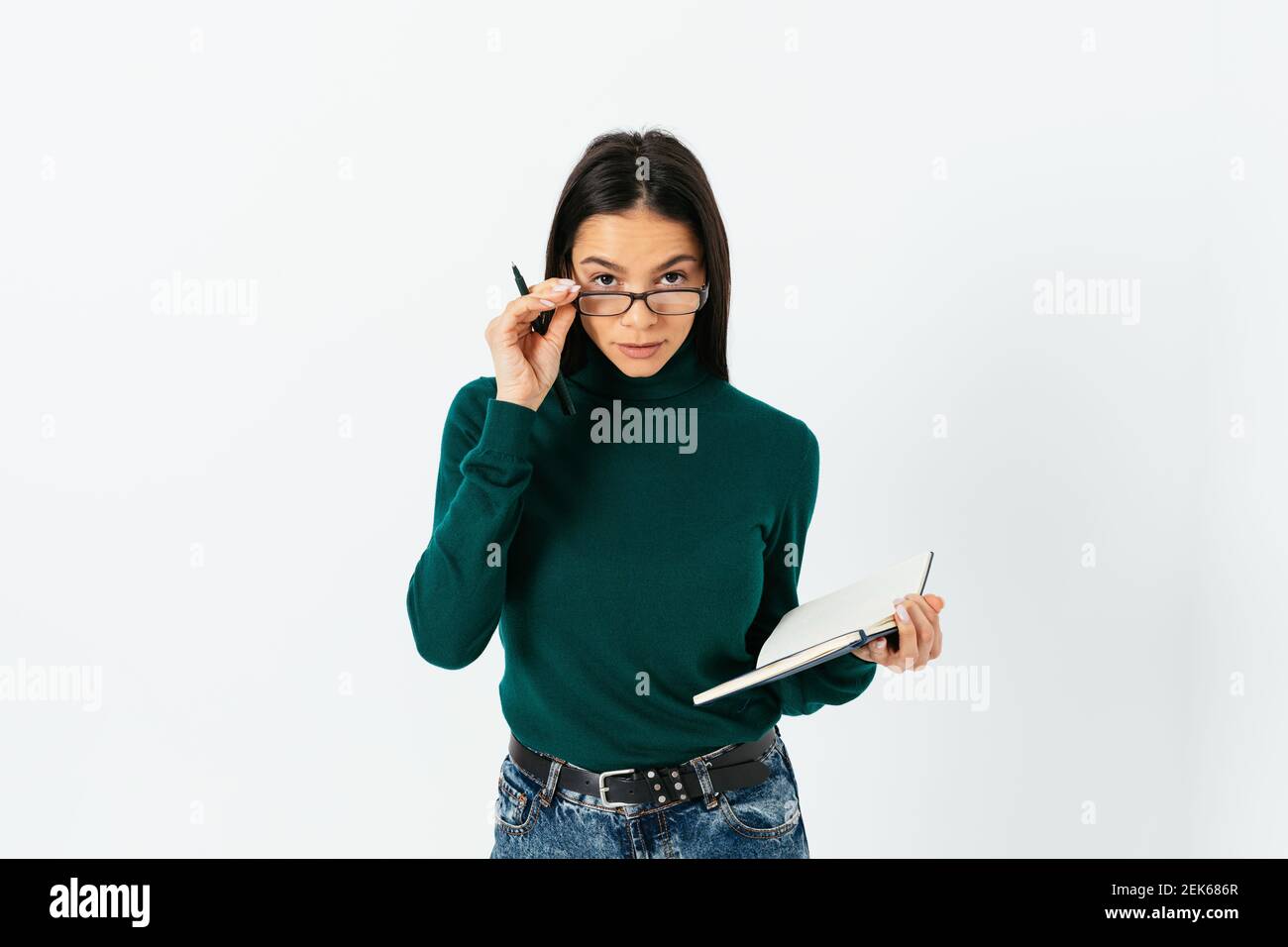 Brunette woman lowered her glasses holding them with her hand looking at camera with calm expression holding pen and notebook on white studio backgrou Stock Photo