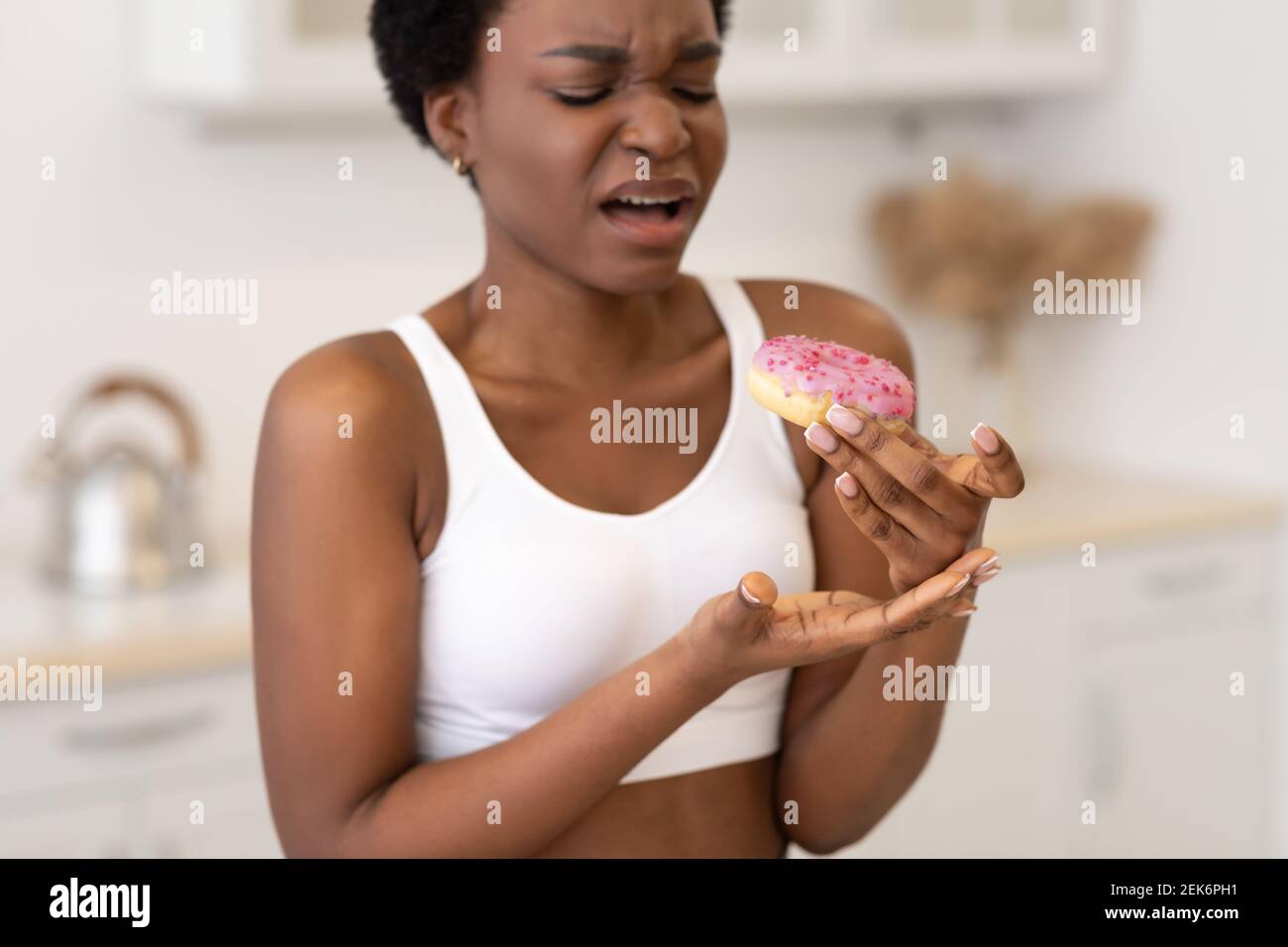 Disgusted African American Woman Eating Unhealthy Donut Standing In Kitchen Stock Photo