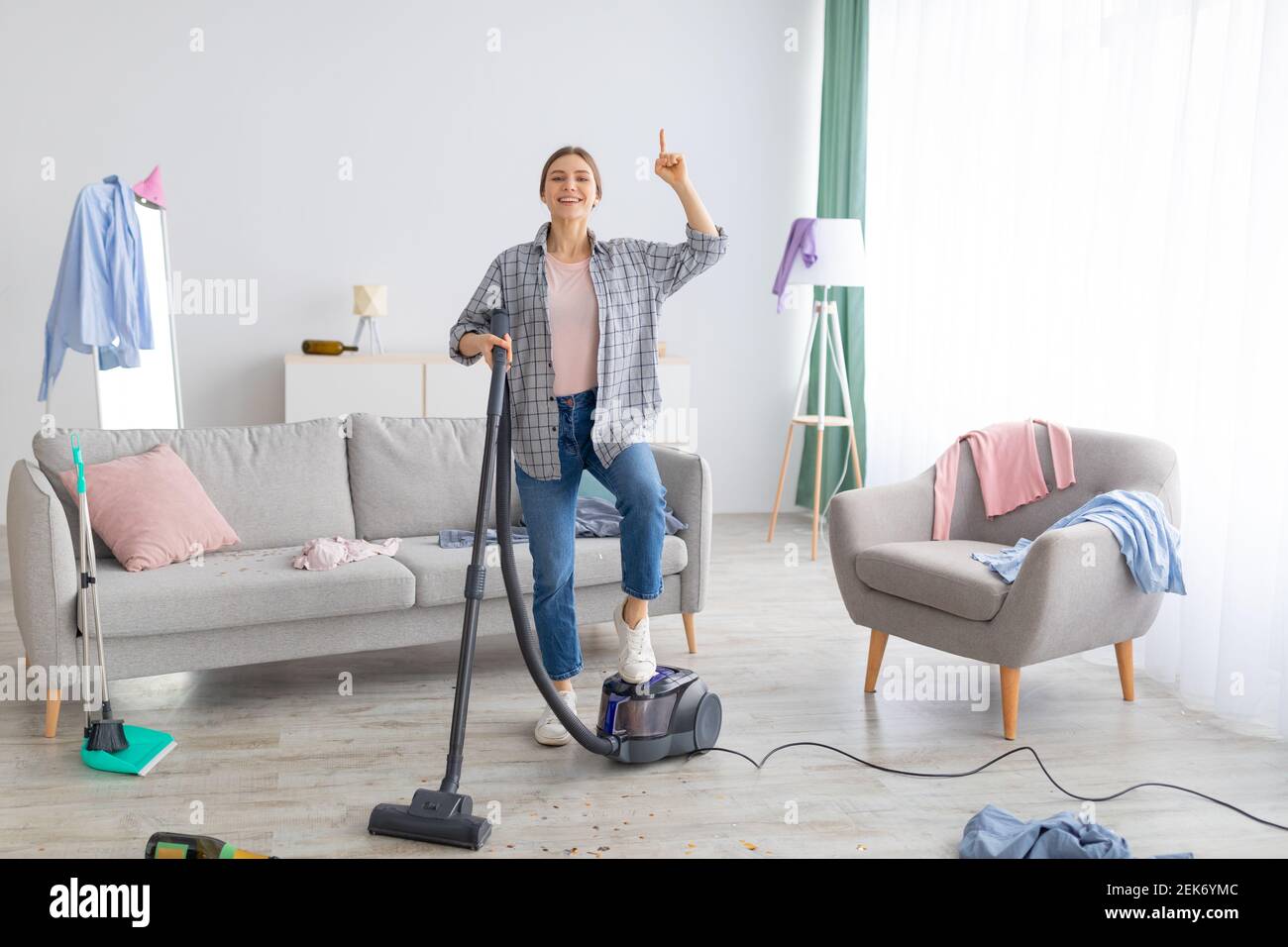 Happy young lady with vacuum cleaner having cleanup idea at messy room after party Stock Photo