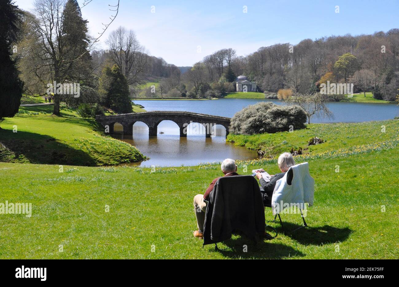 A couple sitting enjoying the spring sunshine while looking across the lake and its Palladian Bridge towards the Pantheon at Stourhead Gardens, Wiltsh Stock Photo