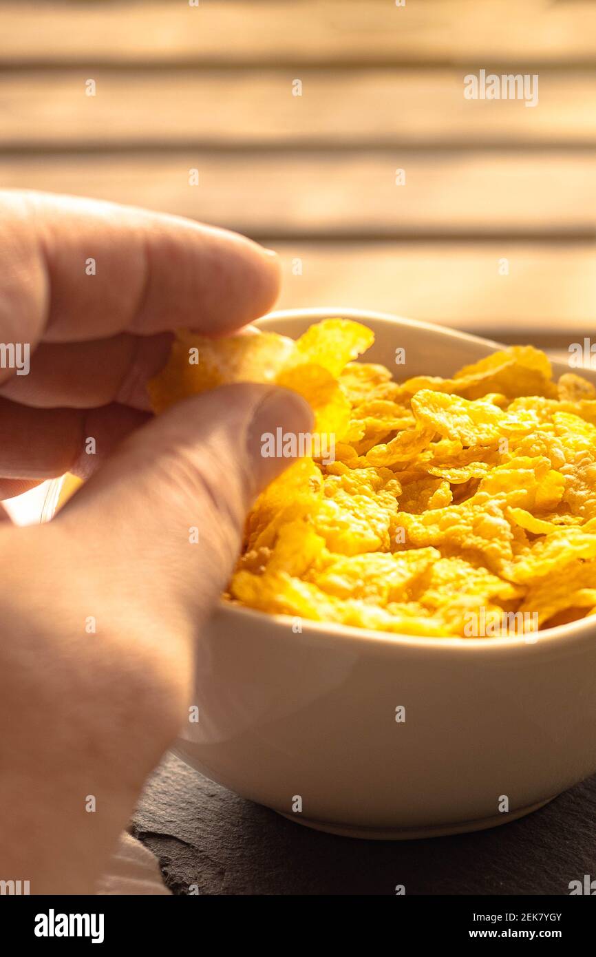 Selective focus of a person picking up cornflakes for breakfast with his hand from a full bowl on a wooden table. Stock Photo