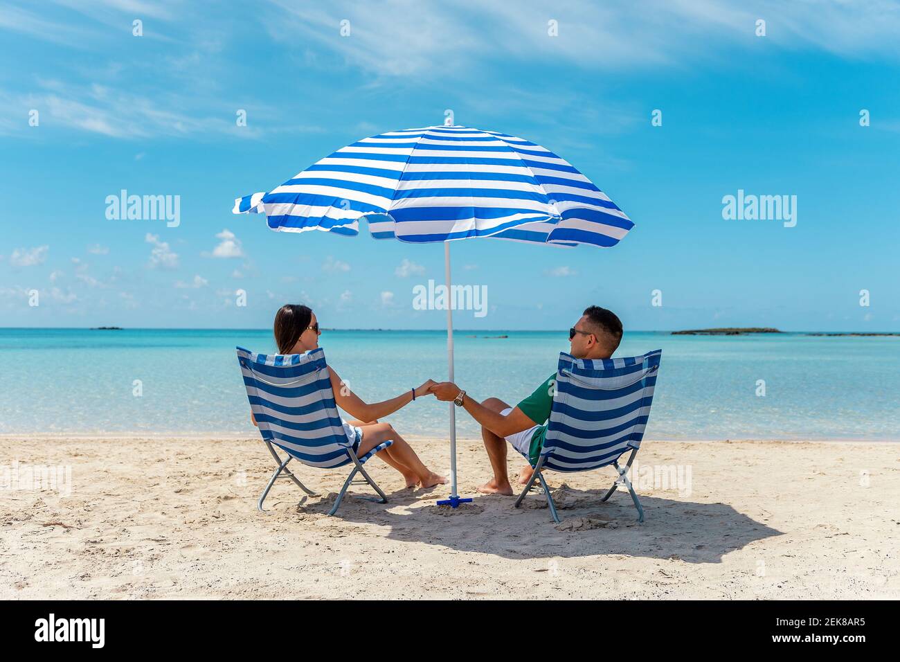 Happy couple is sitting in chairs under parasol on a tropical beach. Summer vacation concept. Stock Photo