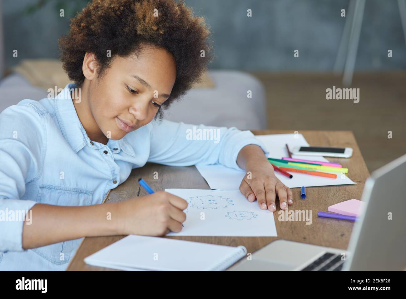 Mixed race teen schoolgirl looking focused while drawing on paper with colorful markers, spending time at home during quarantine Stock Photo