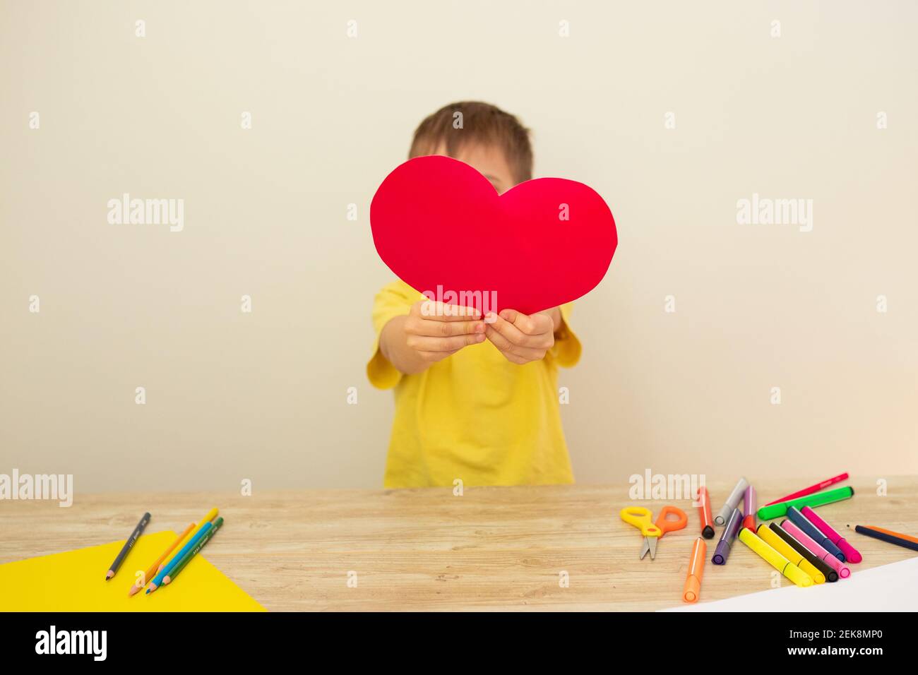 Little boy holding a red heart made of colored paper, covering his face. Concept for March 8, Mother's Day. Postcard Stock Photo