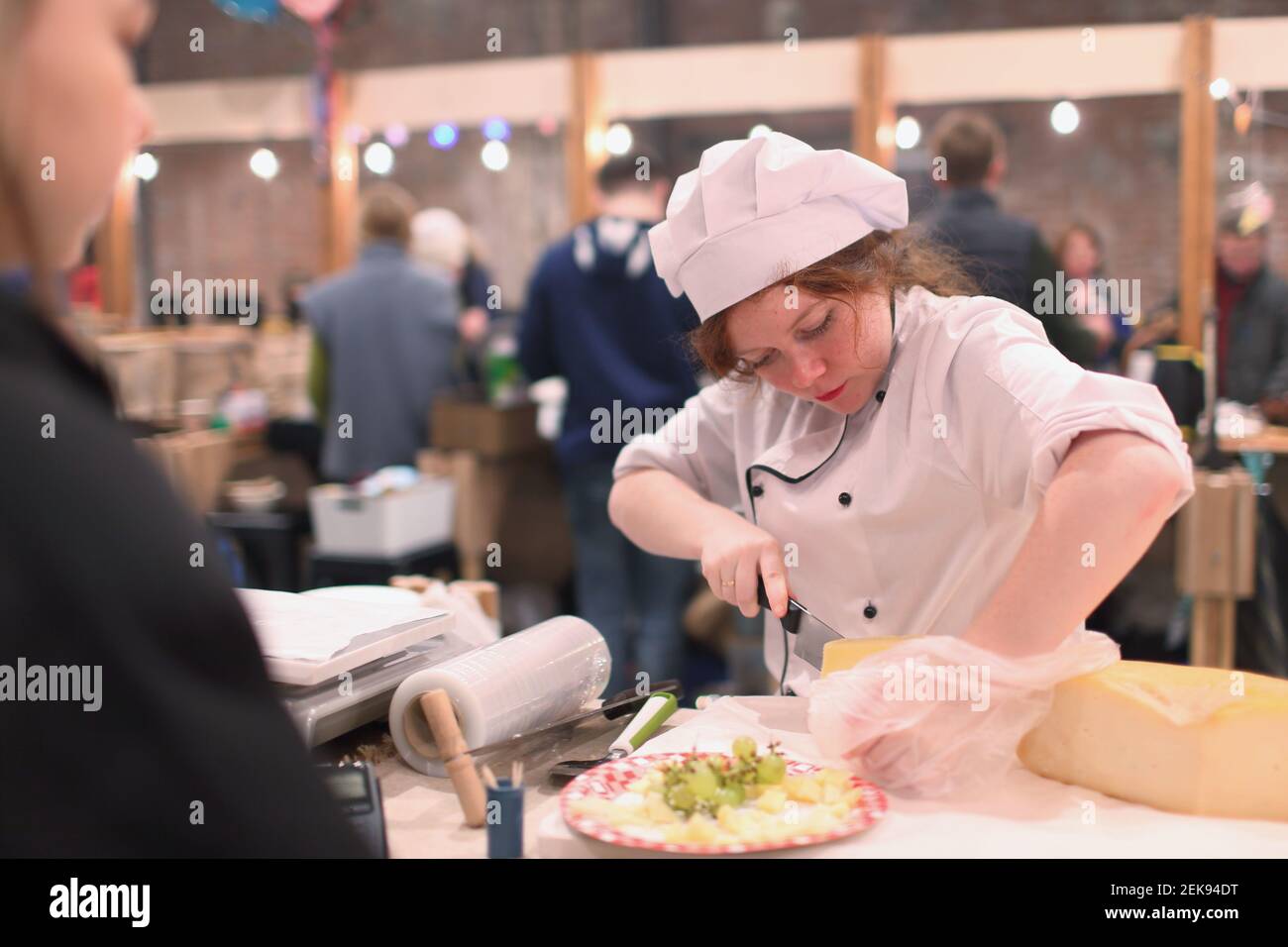 Female market vendor cutting cheese during the fair in Sevkabel public space, St. Petersburg, Russia Stock Photo