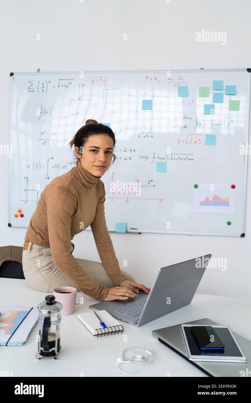 Young female student with laptop sitting on table by whiteboard Stock Photo