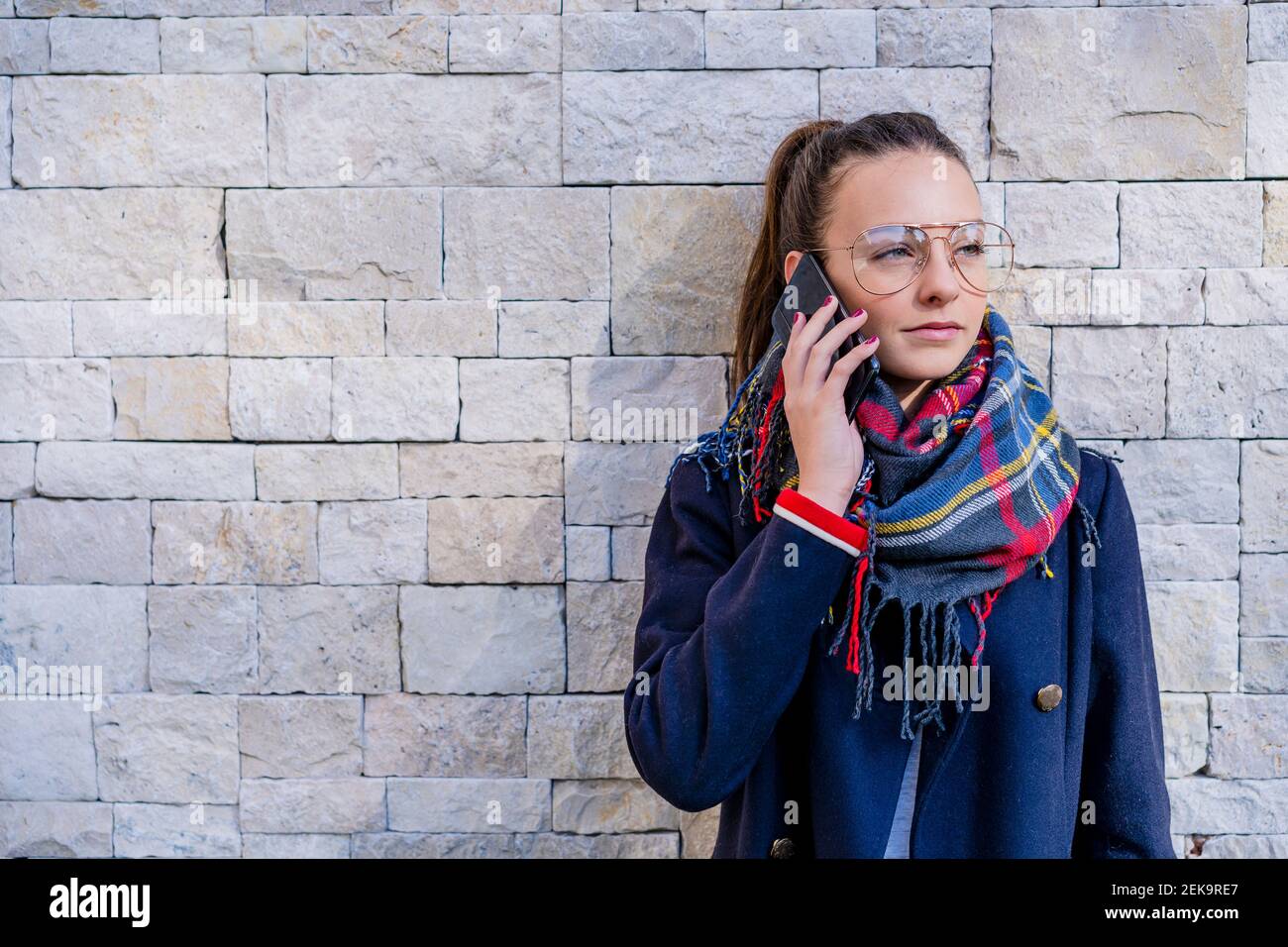 Teenage girl wearing warm clothing talking over smart phone against brick wall at home Stock Photo