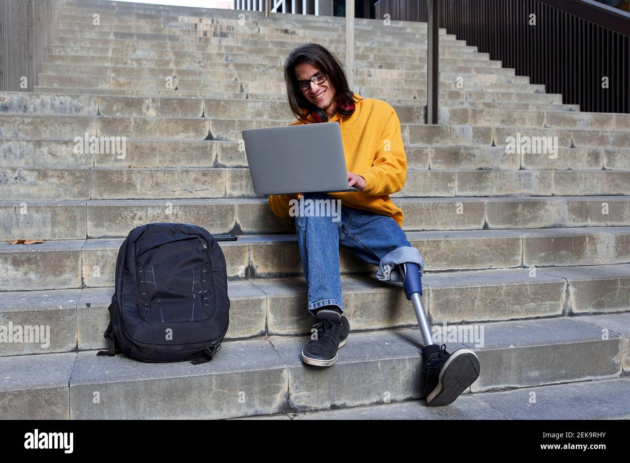 Smiling man with leg prosthesis using laptop while sitting on steps Stock Photo