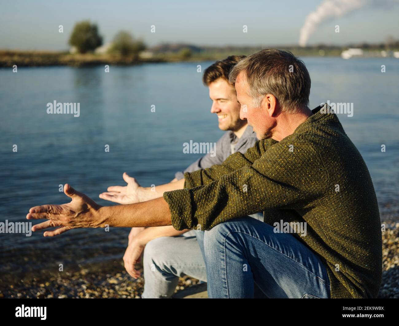 Father gesturing while talking with son by lake on sunny day Stock Photo