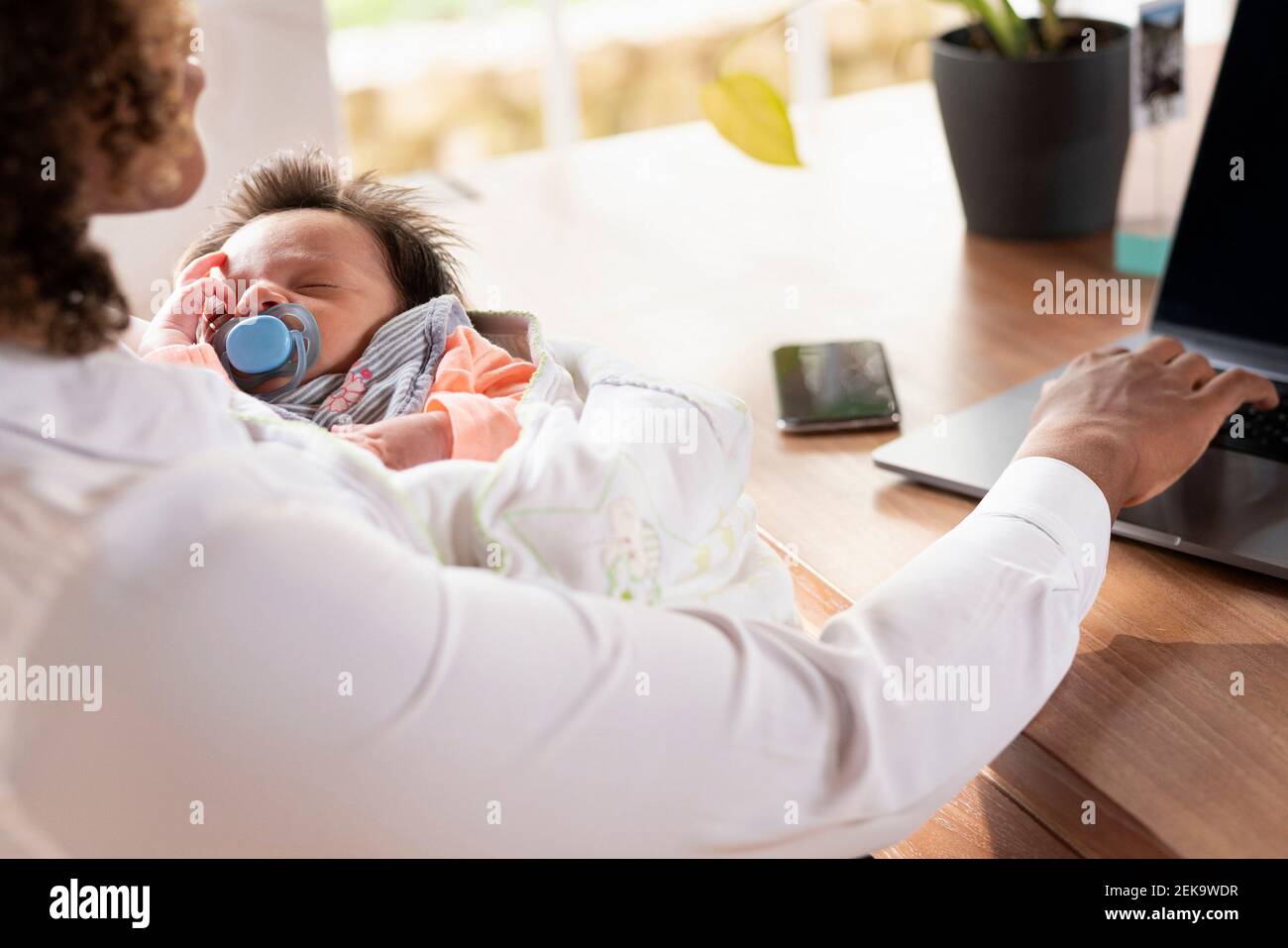 Mother holding sleeping baby while working on laptop at home Stock Photo