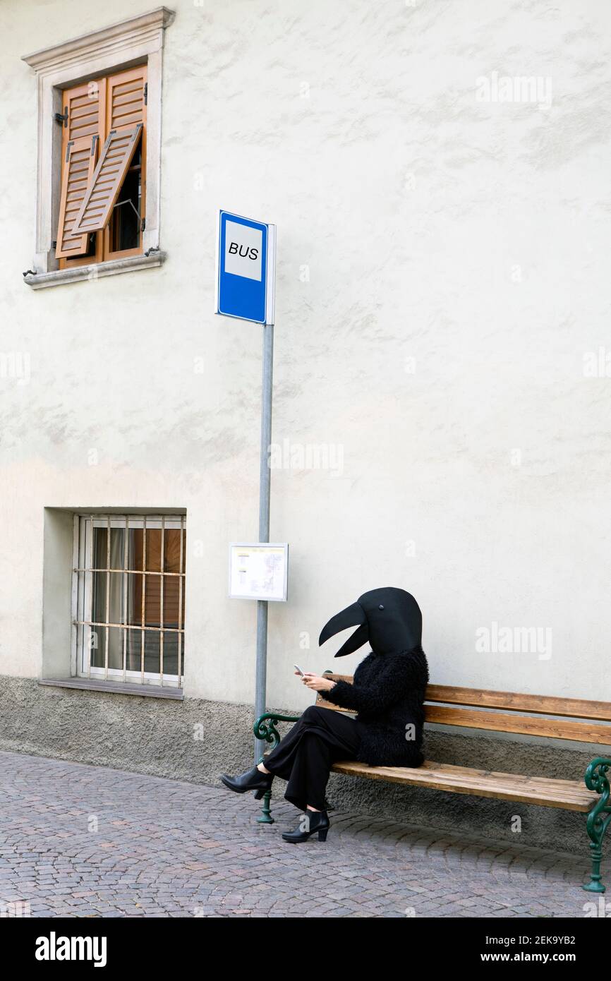 Female in crow costume using mobile phone while sitting on bench of bus stop Stock Photo