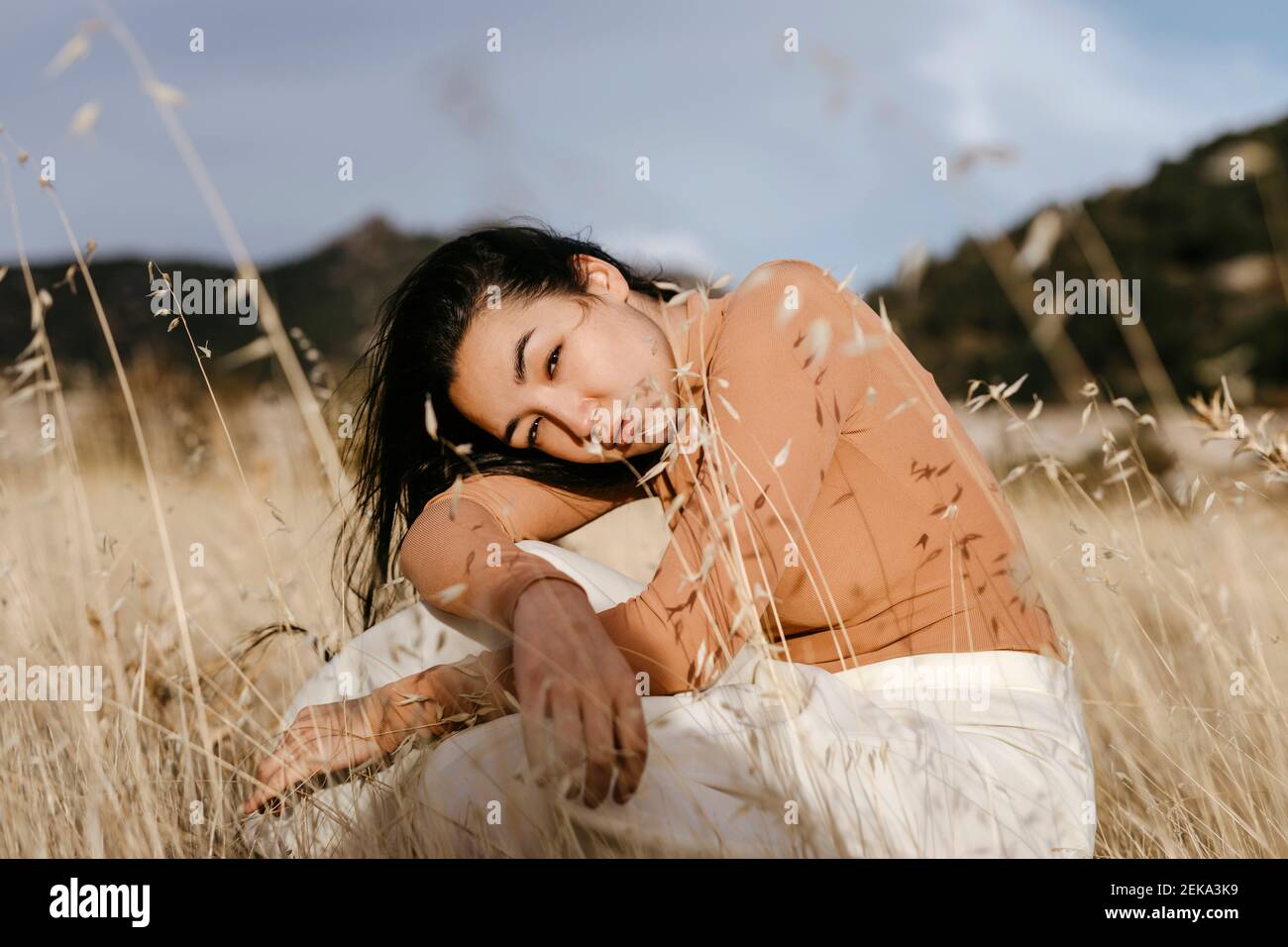 Young woman sitting amidst grass in field against sky during sunset Stock Photo