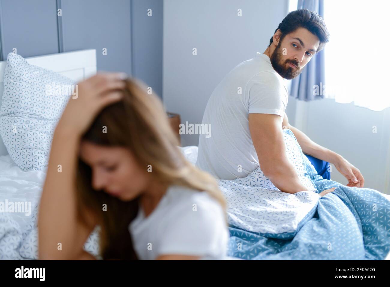 Relationships problems. Young couple sitting in a bedroom and fighting. Both of them looking sad and disappointed. Stock Photo