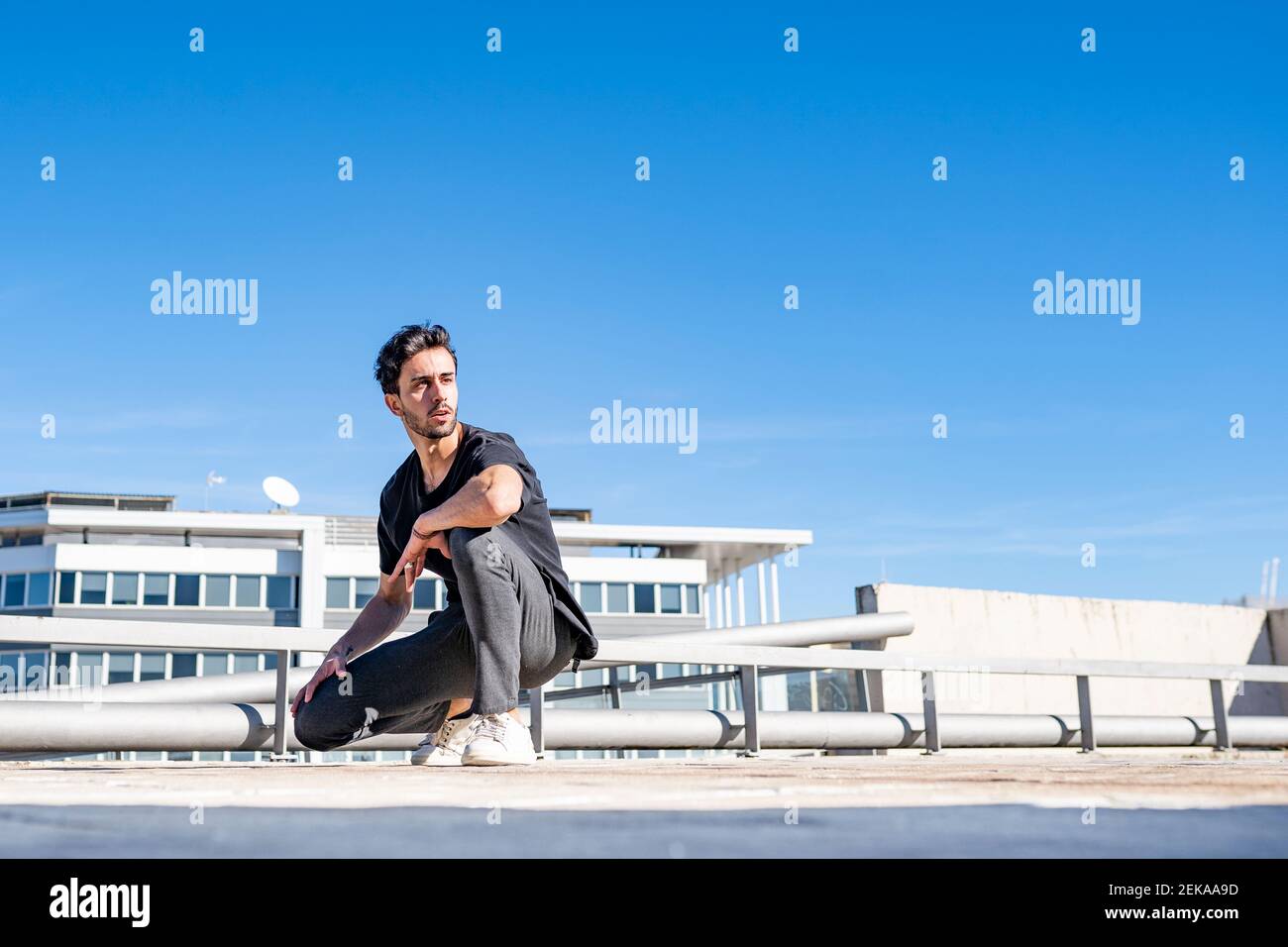 Handsome man looking away while crouching on rooftop during sunny day Stock Photo