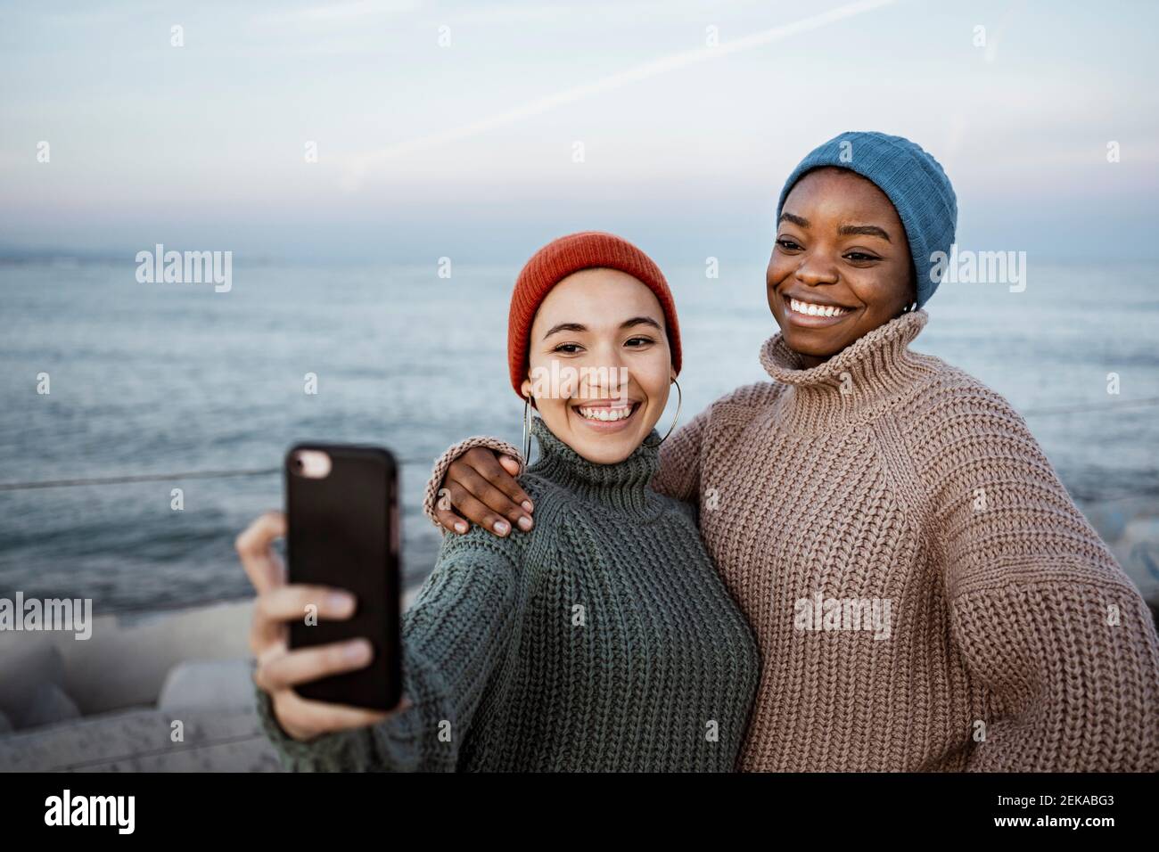 Smiling friends taking selfie through mobile phone while standing against sea Stock Photo