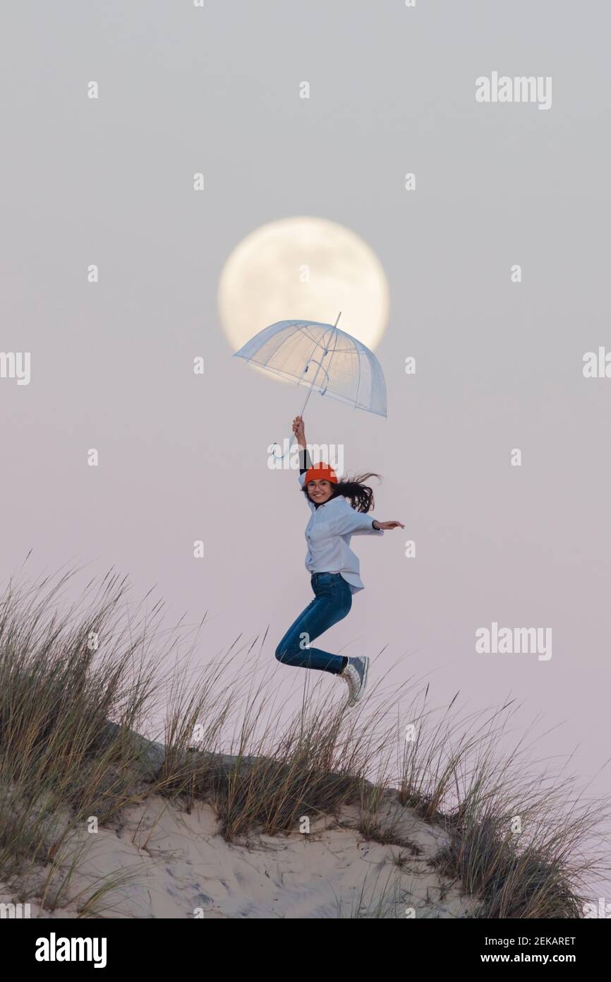 Joyful young woman with umbrella jumping on top of sand dune against sky during dusk Stock Photo