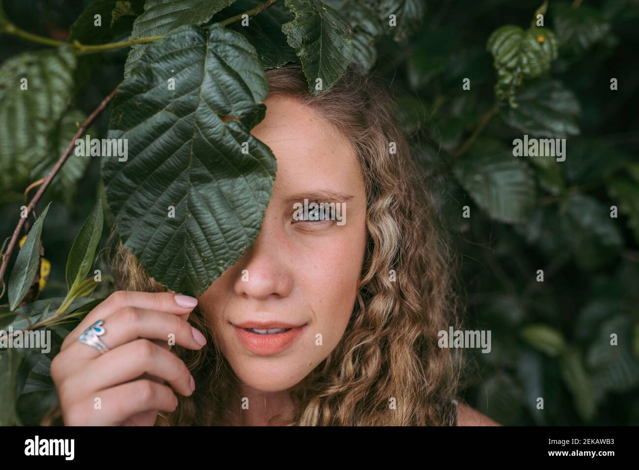 Beautiful blond woman covering eye with leaf in park Stock Photo - Alamy