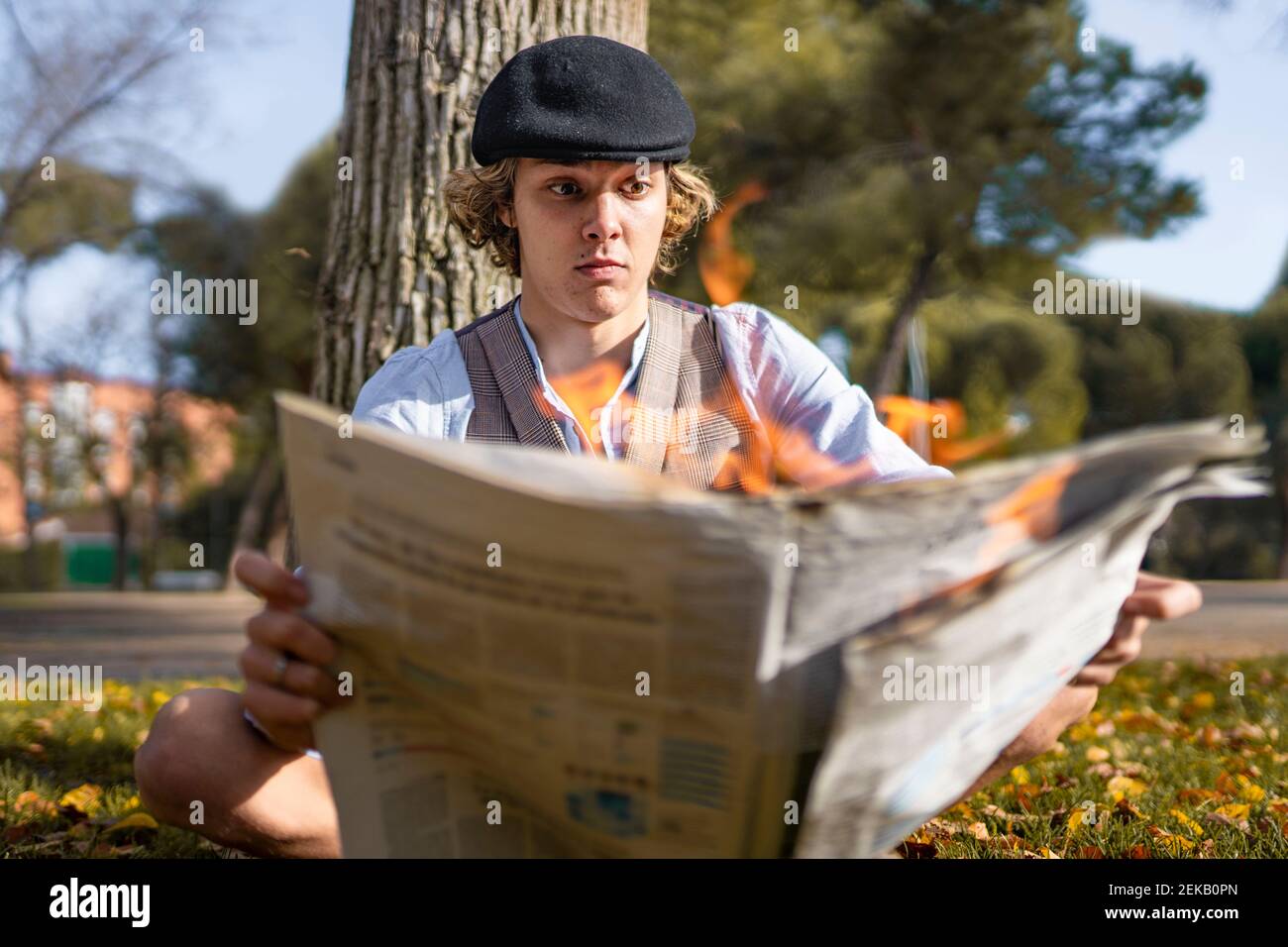 Surprised young man reading lit newspaper while sitting against tree in park Stock Photo