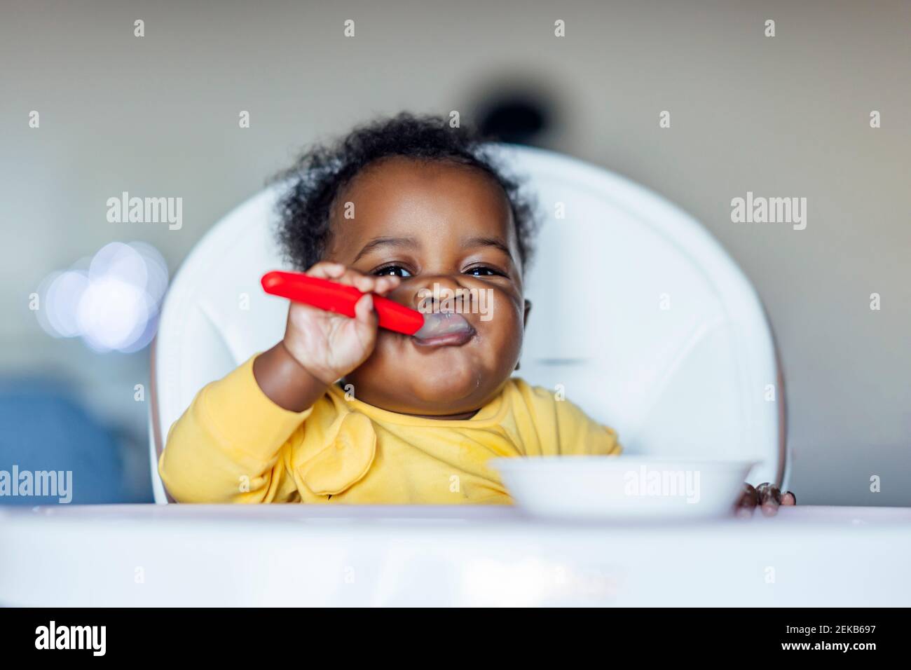 Baby girl eating with spoon while sitting on high chair at home Stock Photo