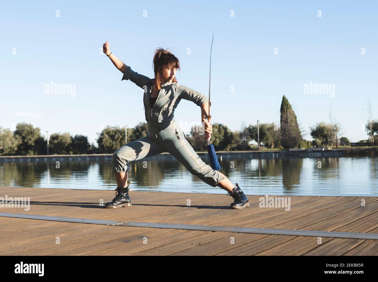 Confident female athlete with sword performing martial arts with dedication by lake on sunny day Stock Photo