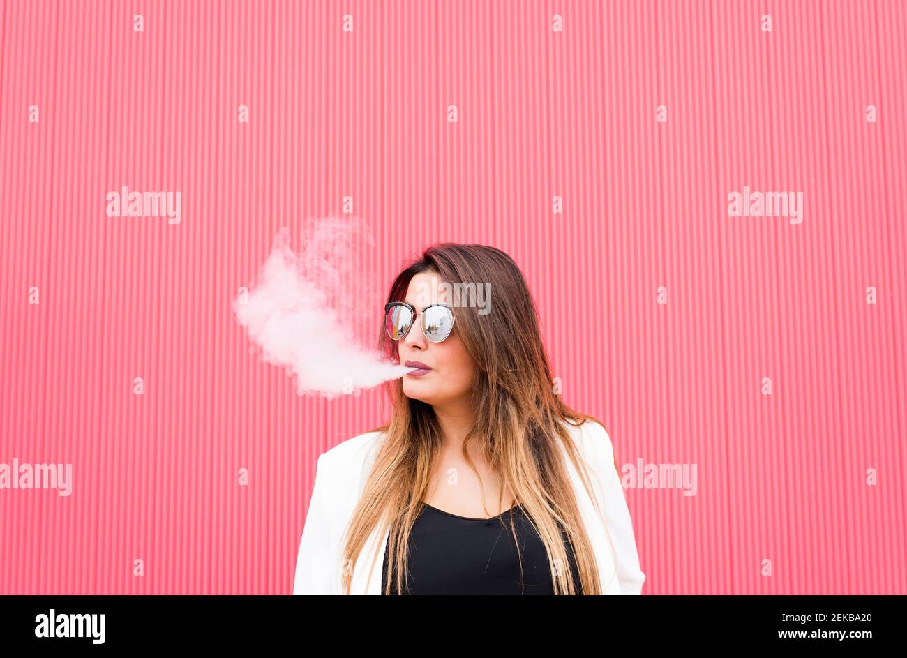 Young woman in sunglasses exhaling smoke against pink wall Stock Photo