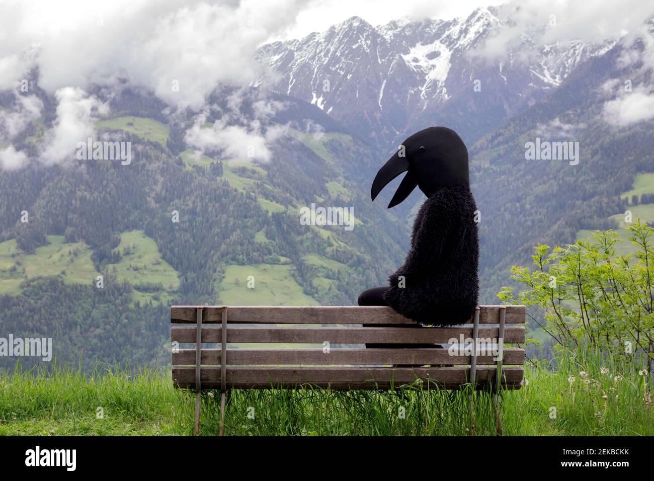 Female in crow costume sitting on bench in front of mountain range Stock Photo