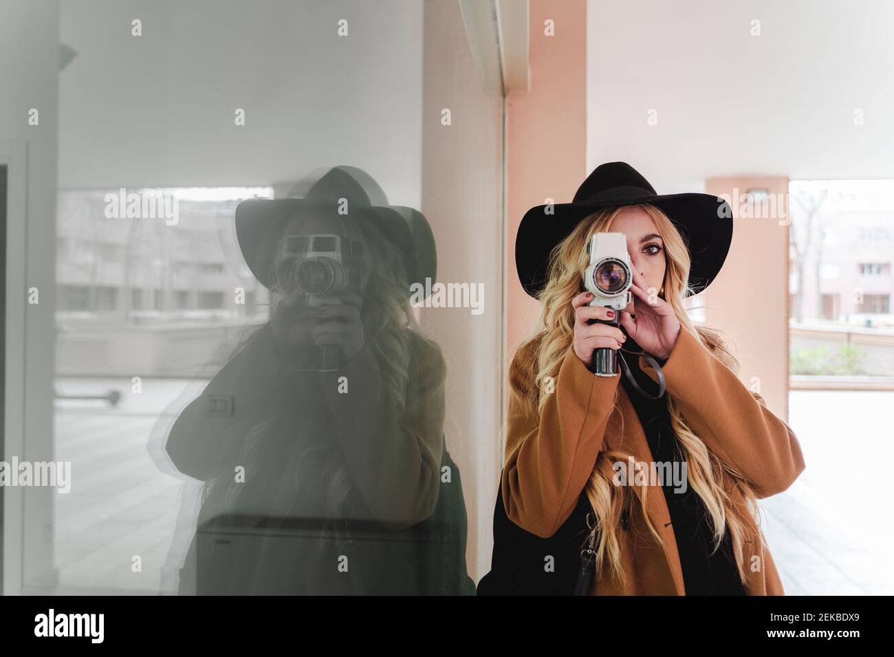 Young woman in hat filming through vintage video camera against reflection in window Stock Photo