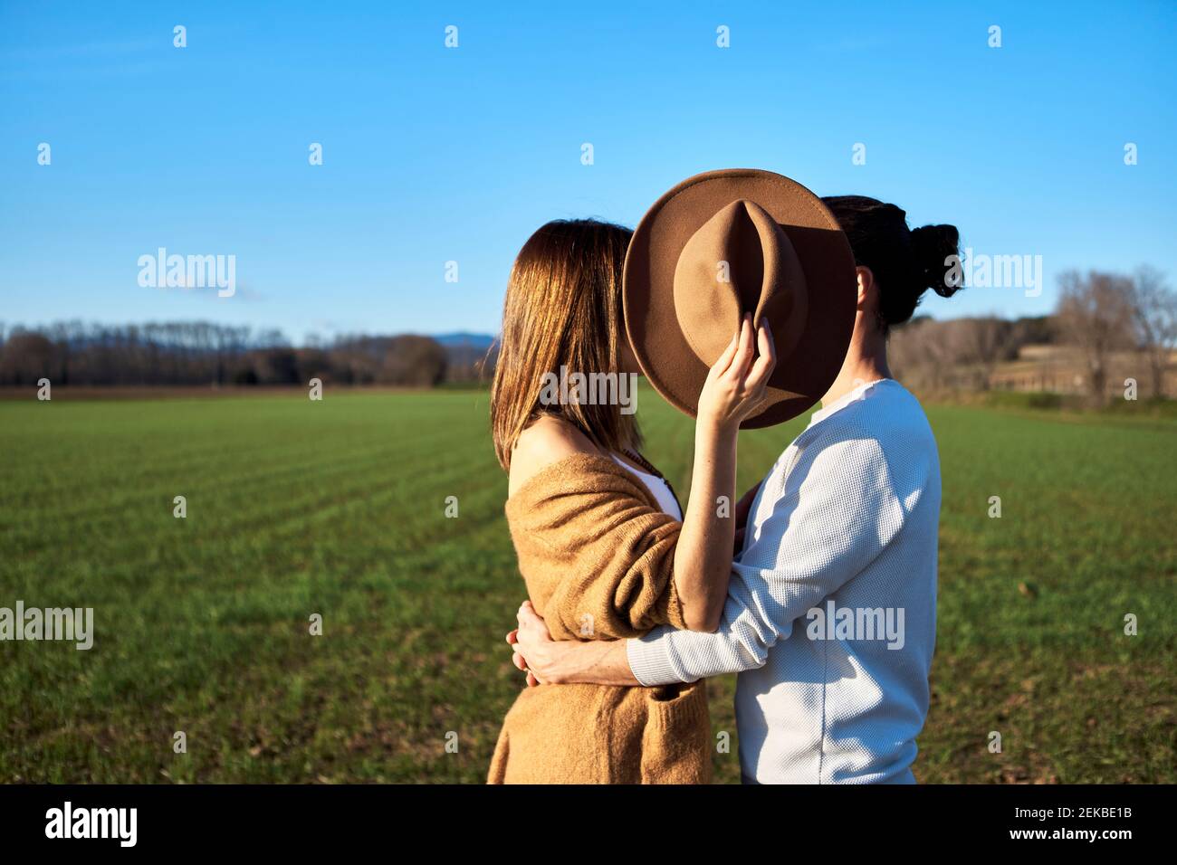 Heterosexual couple embracing while girlfriend covering faces with hat against clear blue sky Stock Photo