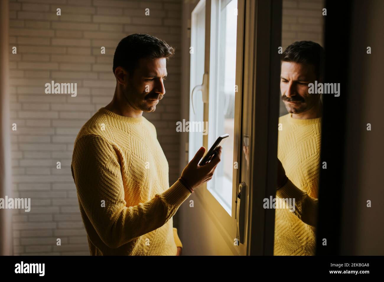 Serious man using smart phone while standing by window at home Stock Photo