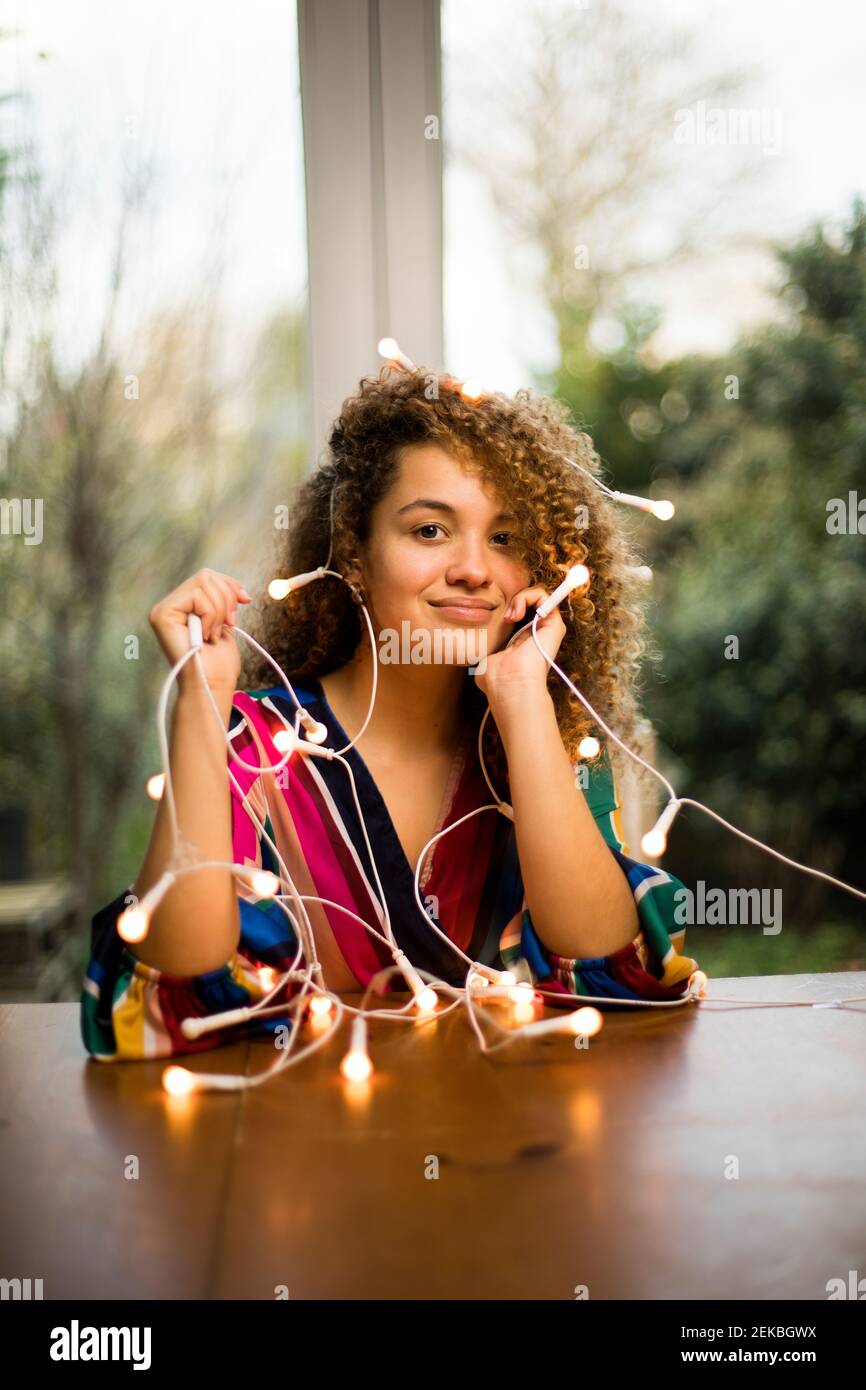 Smiling young woman sitting at table with Christmas lights in living room Stock Photo