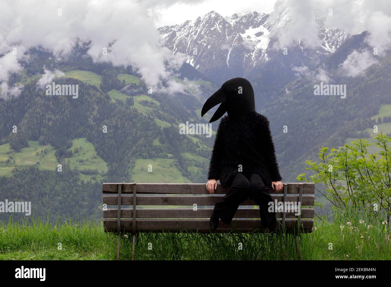 Female in crow costume sitting on bench against mountain range Stock Photo