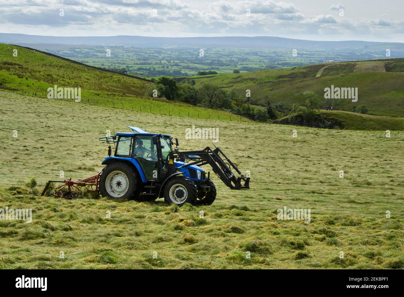 Tractor haymaking (pulling & towing tedder, tedding farm silage crop) in scenic farmland pasture field near Grassington, Yorkshire Dales, England, UK. Stock Photo
