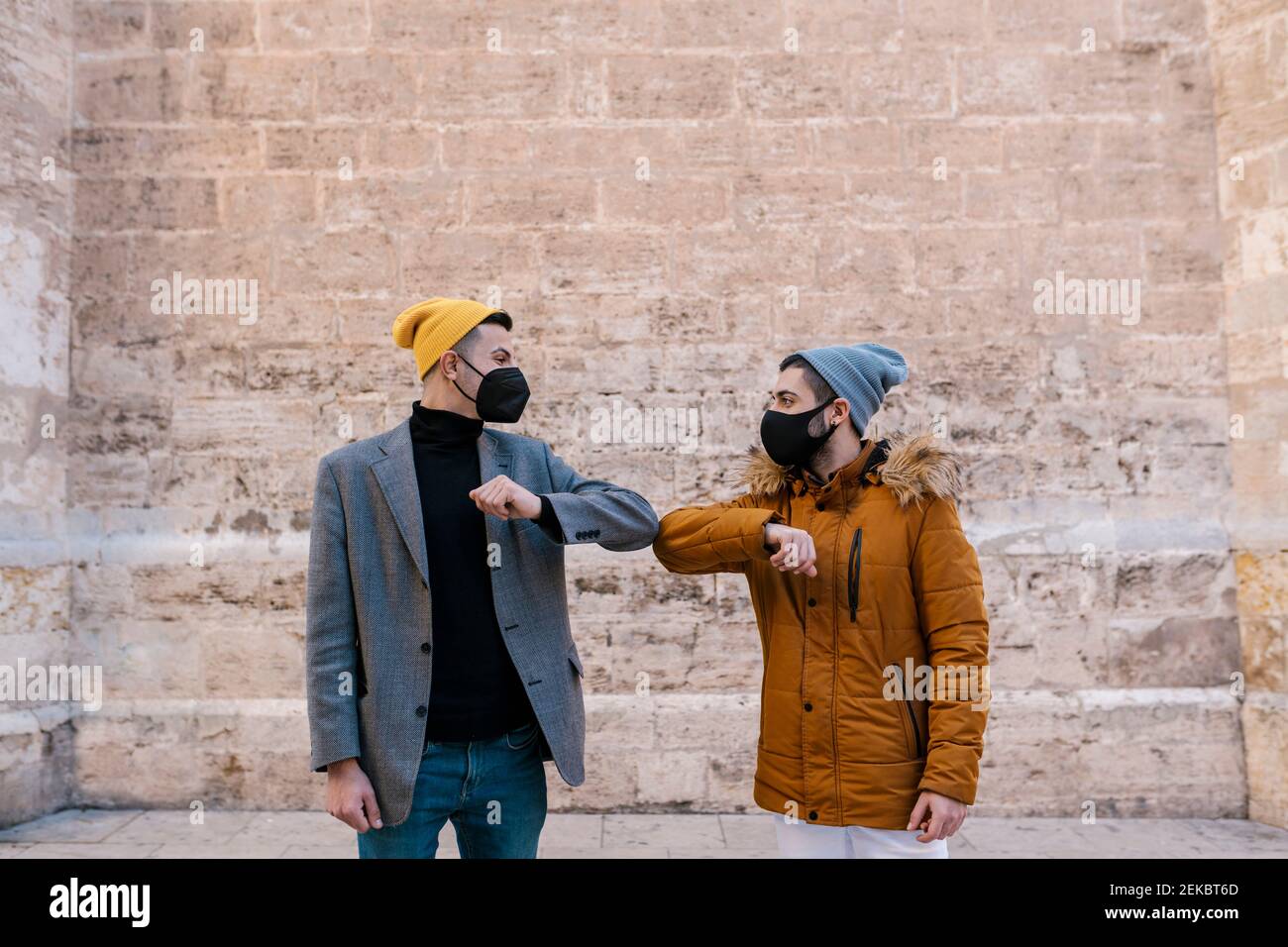 Young men wearing knit hat and protective face mask giving elbow bump while greeting against wall Stock Photo