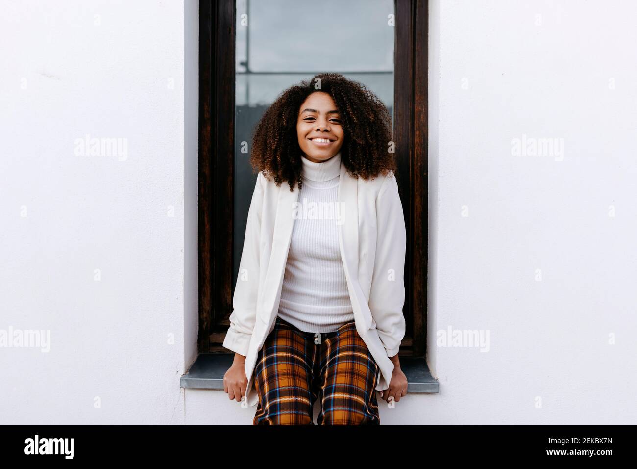Smiling young woman with afro hair sitting on window sill Stock Photo ...
