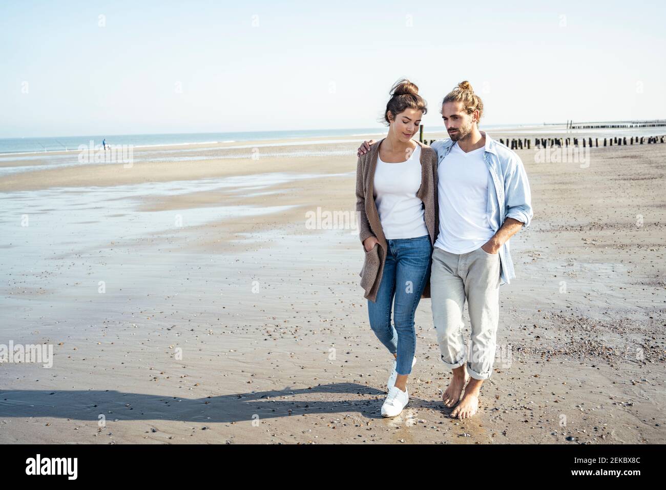 Young couple with hands in pockets taking walk on beach during sunny day Stock Photo